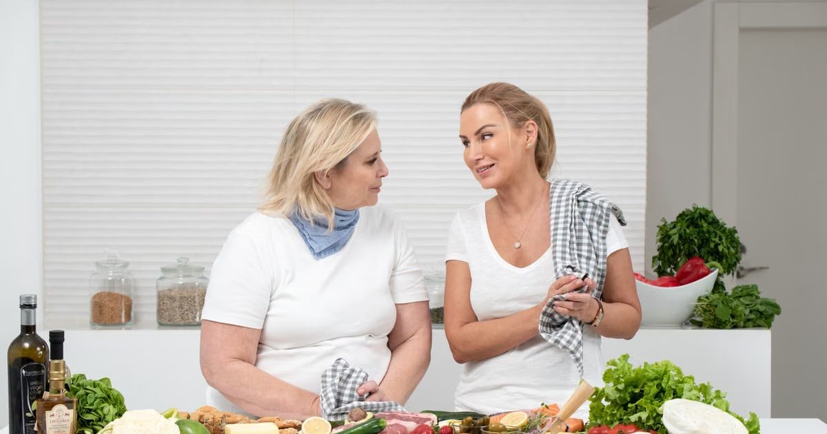 Two women in a kitchen preparing fresh vegetables and cheese with olive oil and spices.