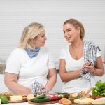 Two women in a kitchen preparing fresh vegetables and cheese with olive oil and spices.