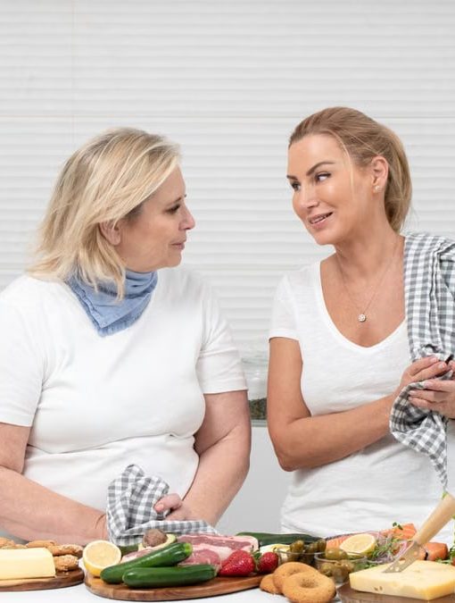 Two women in a kitchen preparing fresh vegetables and cheese with olive oil and spices.
